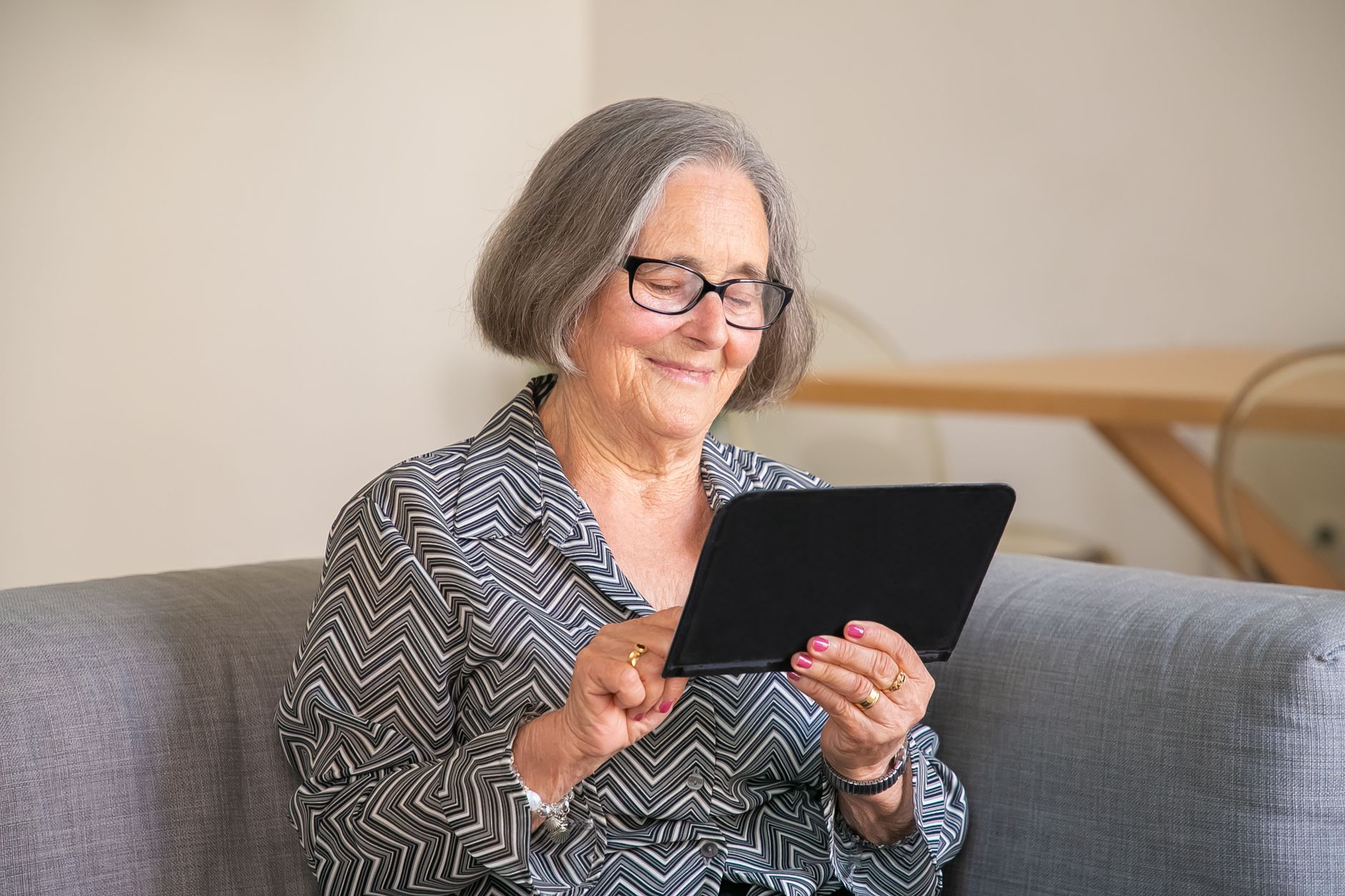 Femme aux cheveux gris et aux lunettes noires, souriante, qui utilise une tablette assise sur un canapé gris